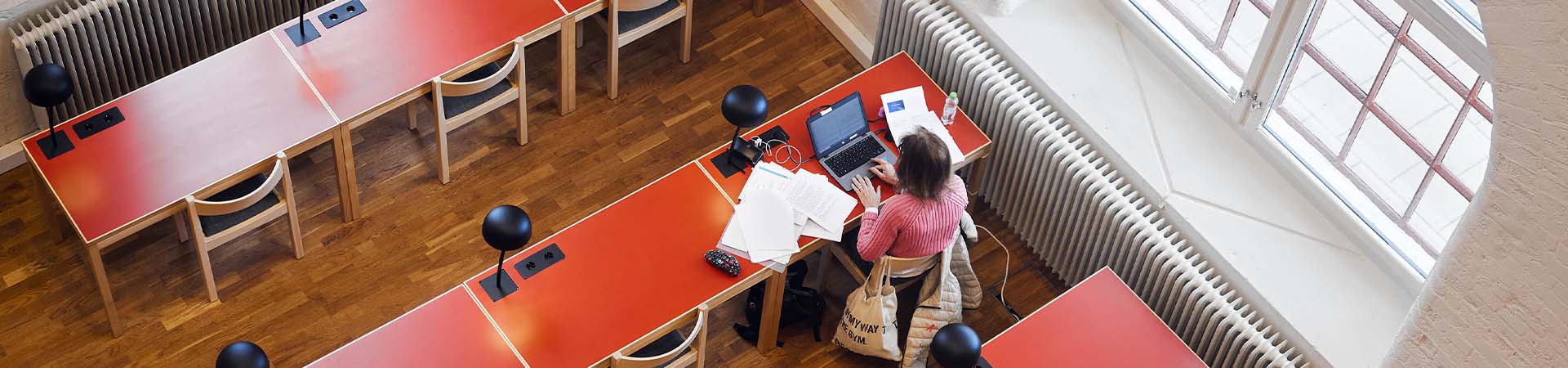 En ensam student sitter och studerar med en laptop och en massa papper utspridda på det röda bordet. Bordet är intill ett stort fönster. Bilden är tagen uppifrån.