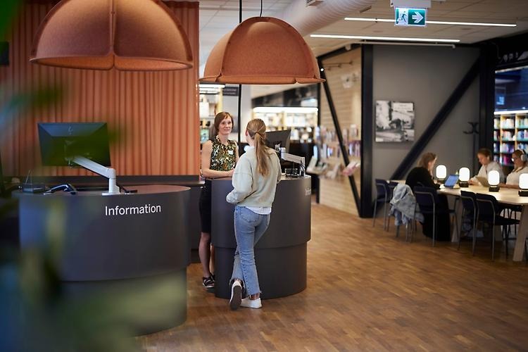 A librarian is standing next to the information desk, talking to a student. Behind the desk, you can se a table with students who are studying.